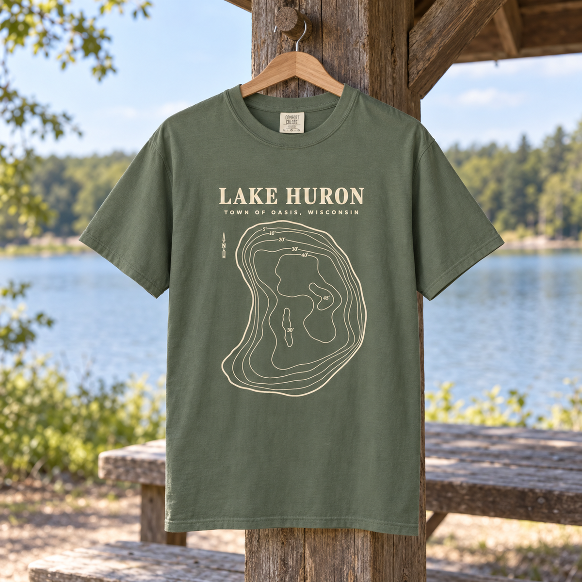 Adult Moss — product lifestyle mockup of the Lake Huron Field Survey T-shirt with cream contour-map artwork and Town of Oasis, Wisconsin lettering, in a generated wooded lakeside setting.