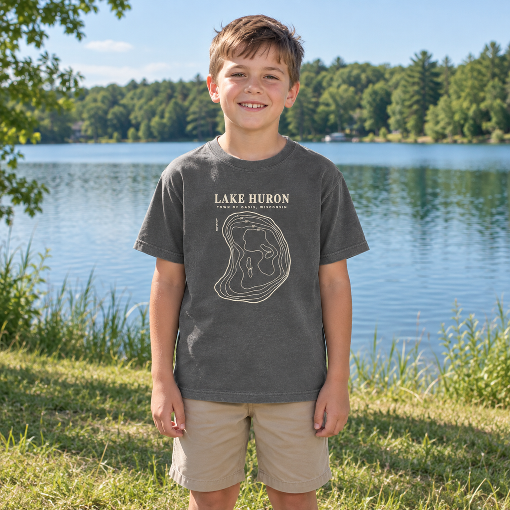 Kids Pepper — lakeshore lifestyle mockup of the Lake Huron Field Survey T-shirt with cream contour-map artwork and Town of Oasis, Wisconsin lettering, in a generated wooded lakeside setting.