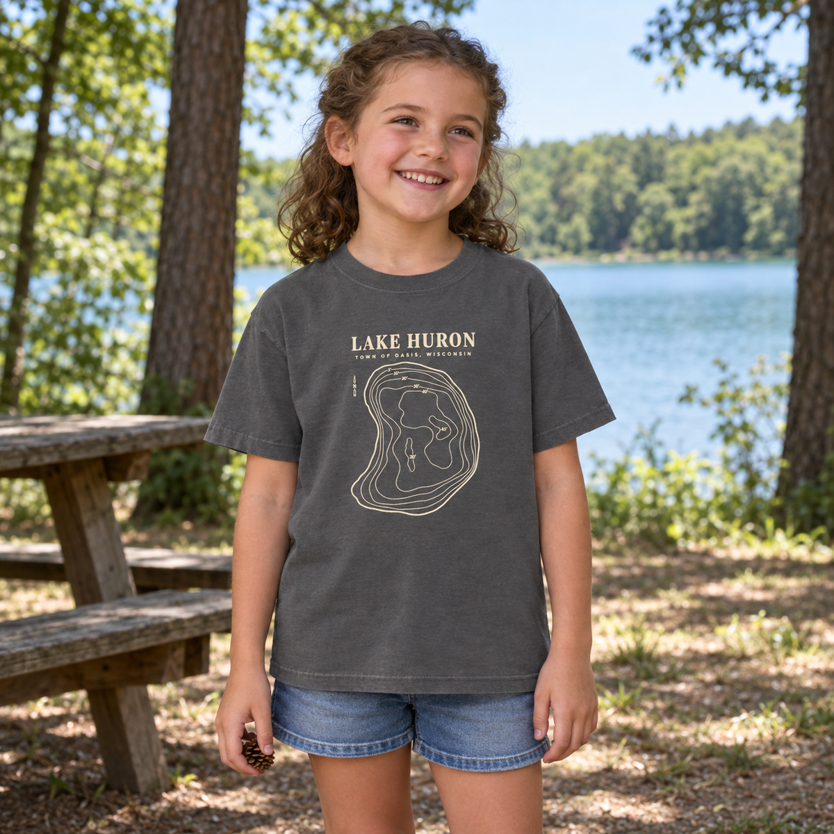 Kids Pepper — park lifestyle mockup of the Lake Huron Field Survey T-shirt with cream contour-map artwork and Town of Oasis, Wisconsin lettering, in a generated wooded lakeside setting.