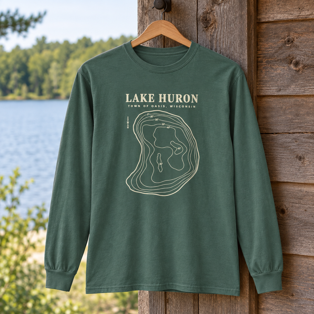 Blue Spruce Comfort Colors 6014 adult Lake Huron long-sleeve shirt with cream contour artwork and Town of Oasis, Wisconsin lettering, hanging at a cabin beside a small Wisconsin lake in a generated lifestyle scene.