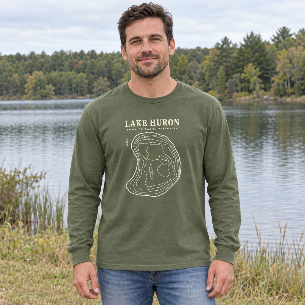 Hemp Comfort Colors 6014 adult Lake Huron long-sleeve shirt with cream contour artwork and Town of Oasis, Wisconsin lettering, worn by an adult beside a small Wisconsin lake in a generated lifestyle scene.